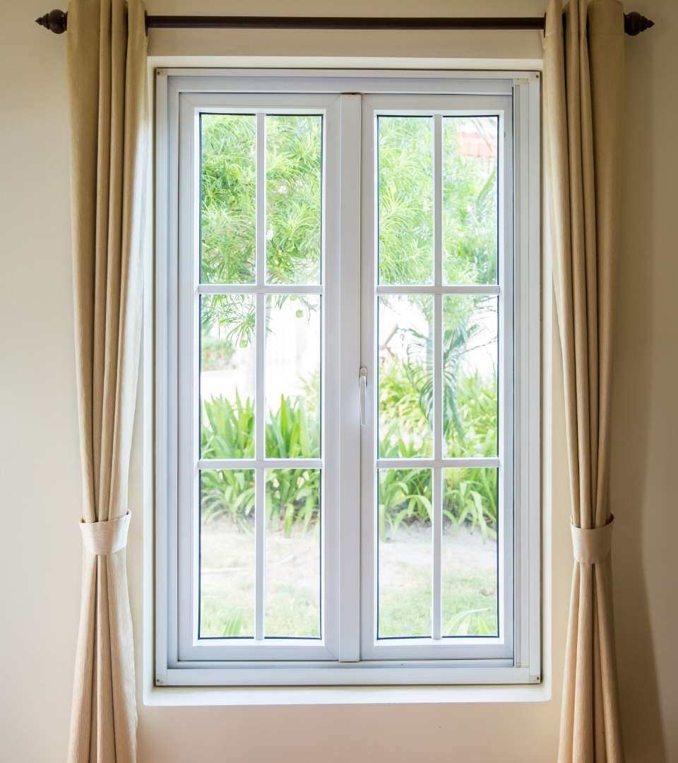 This picture is taken from inside a residential or commercial property and is looking through a beautiful glass window. The window has a white frame and tan curtains. Pane Free Glass in Manchester, NH, specializes in window installations that will conserve energy and save you money.